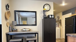 Een kleine keuken met een donker houten kastje, een inbouwmagnetron en een koelkast. Er staat een klein tafeltje met twee krukken bij, en erboven hangt een spiegel.