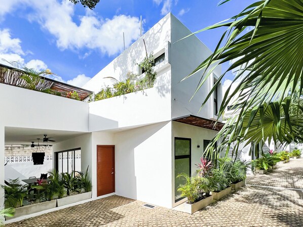 A modern two-story house with a flat roof, large windows, and a covered patio area with potted plants.