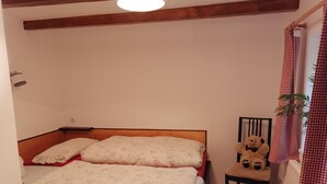 Une chambre avec une tête de lit en bois, un lit recouvert d’une couette à motifs floraux, une chaise sur laquelle est posé un ours en peluche, et une fenêtre ornée de rideaux à carreaux.
