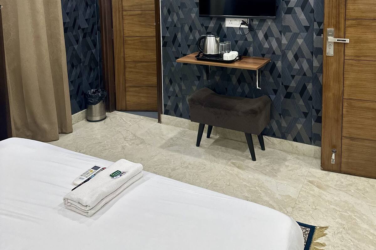 A hotel room with a bed, a TV mounted on the wall, a small table with a kettle, and a trash bin.