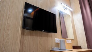 Televisão de tela plana fixada numa parede de madeira, uma pequena mesa com abajur e uma tomada.