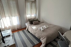 Een slaapkamer met een bed, kussens, een nachtkastje, een stoel en een raam met gordijnen.