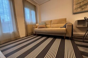 Quarto com sofá, tapete listrado, janela com cortinas e prateleira na parede.