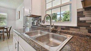 A kitchen with a double sink, granite countertop, white cabinets, and a window with a view of greenery. - Charming Dade City Home with Fire Pit & Garden (Dade City)