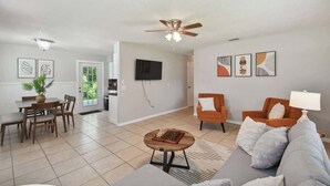 A living room with a sofa, armchairs, a coffee table, a dining table with chairs, a TV mounted on the wall, and a ceiling fan. - Charming Dade City Home with Fire Pit & Garden (Dade City)