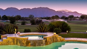 A swimming pool with a stone border, surrounded by a landscaped garden with various plants and a view of a golf course and mountains in the background.