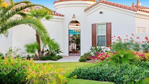 A white building with a tiled roof, a central archway, and a small garden with various plants and flowers.