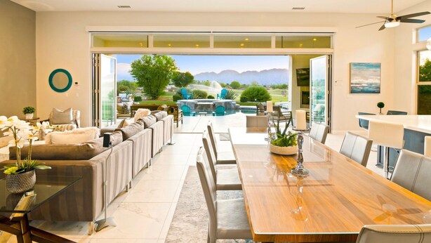 A spacious dining area with a long wooden table, comfortable seating, and a view of a landscaped garden and mountains.