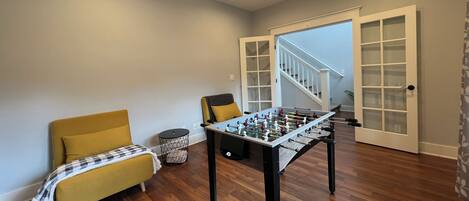 A room with a foosball table, a yellow sofa, a ceiling fan, and a staircase.