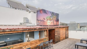 A rooftop bar with a sunset mural, wooden bar counter, and outdoor seating. - Happy Day Hotel (Praia)