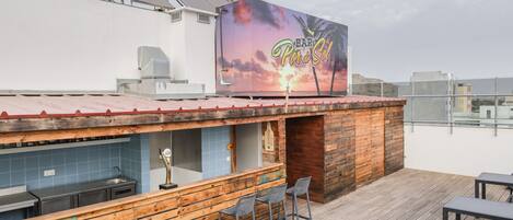 Un bar en la azotea con un mural del atardecer, barra de madera y mesas al aire libre.
