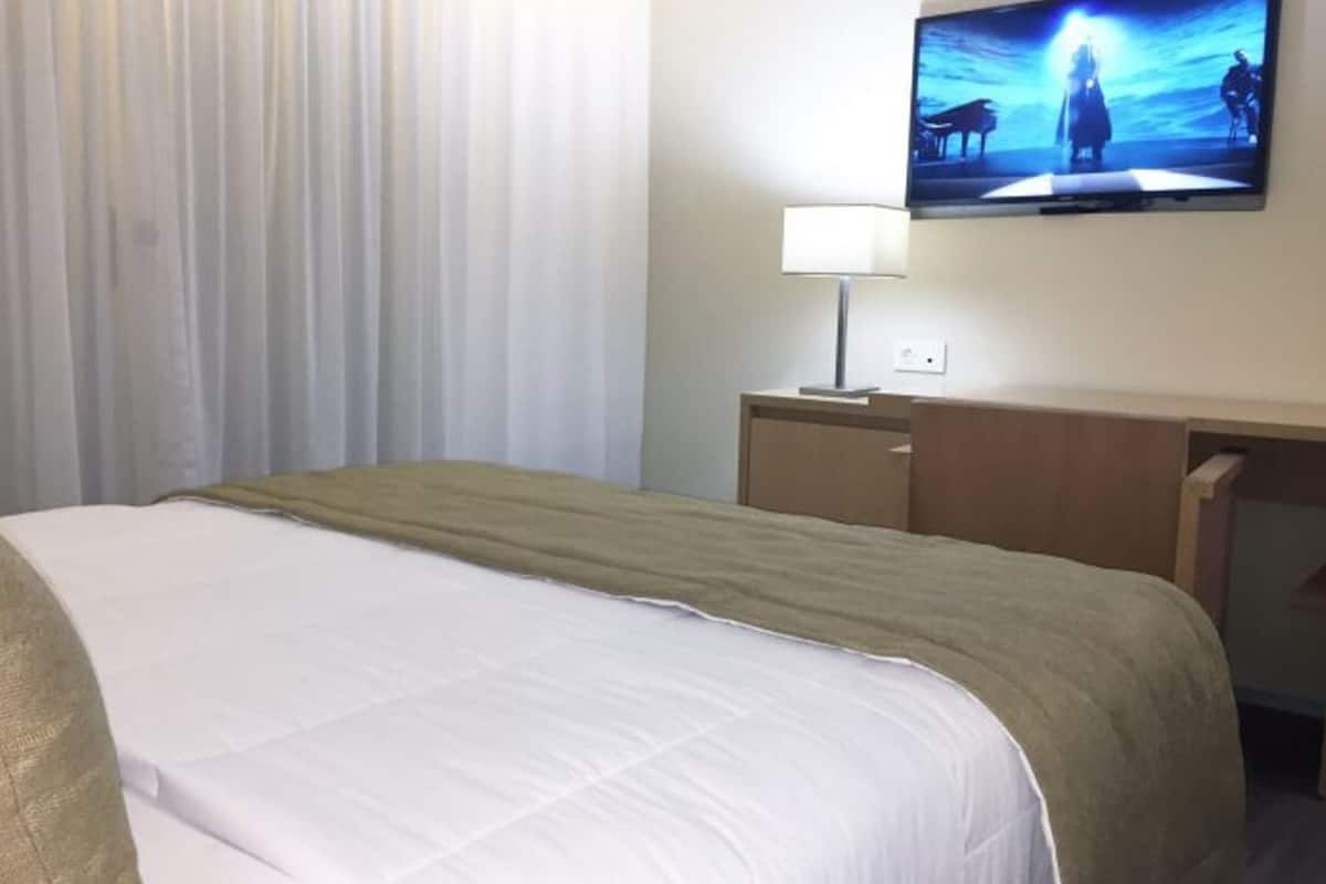 A hotel room with a bed, a TV mounted on the wall, a desk, and a lamp.