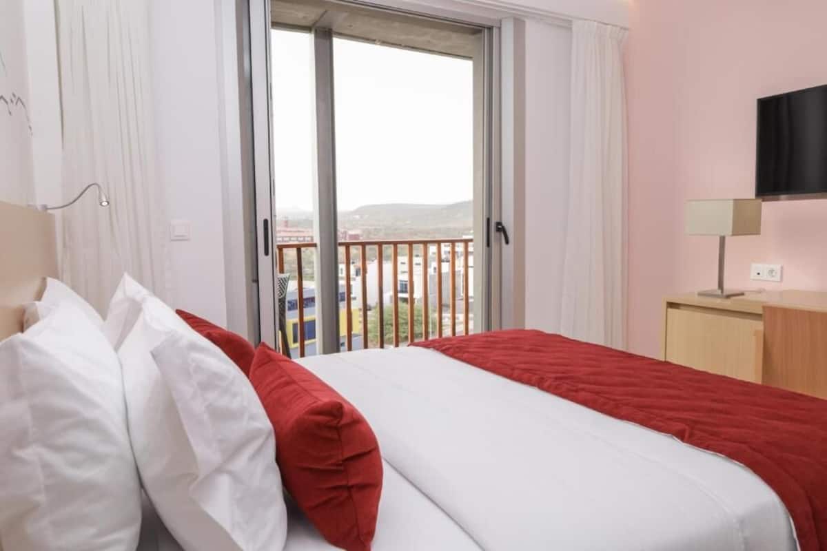 A hotel room with a bed, a TV, a desk, and a window with a view of a building and a balcony.