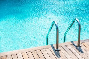 Terraza de madera con barandillas metálicas junto a una piscina azul cristalina.