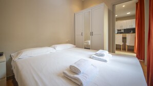 A neatly made bed with white linens and towels, a white wardrobe, and a view into a modern kitchen area. - 2 bed apartment in Barceloneta steps from the beach (Barcelona)