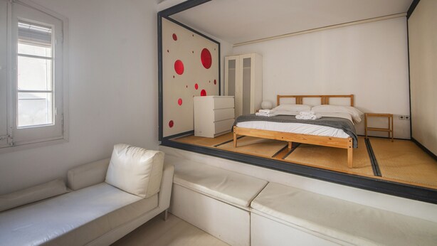 A bedroom with a bed, a white sofa, a wardrobe, and a large mirror reflecting the room.