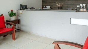 Un área de recepción con un mostrador blanco, dos lámparas colgantes, una planta en maceta y una silla roja.