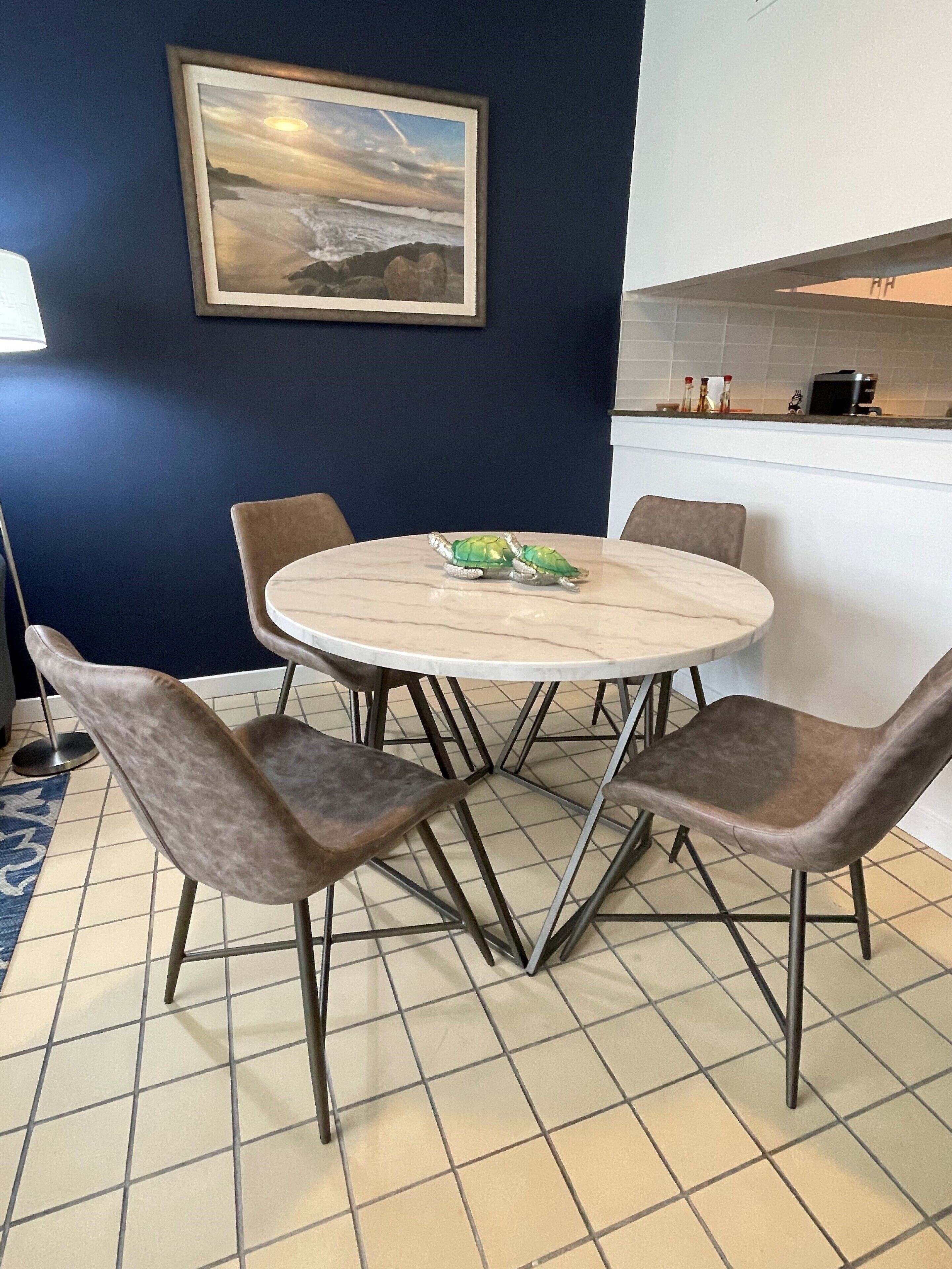A dining area with a round table and four chairs, a painting on the wall, and a kitchen counter with appliances.