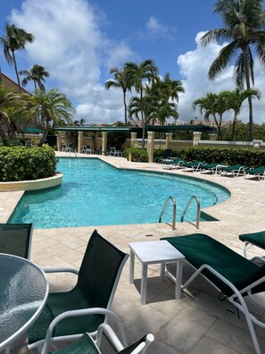 Une piscine entourée de palmiers et d’un bâtiment en arrière-plan, avec des chaises longues et une table à proximité.