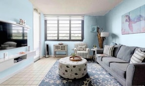 A living room with a sofa, a round ottoman, a television, and a window with blinds. - Carey Beach Villa at Palmas del Mar (Palmas del Mar)