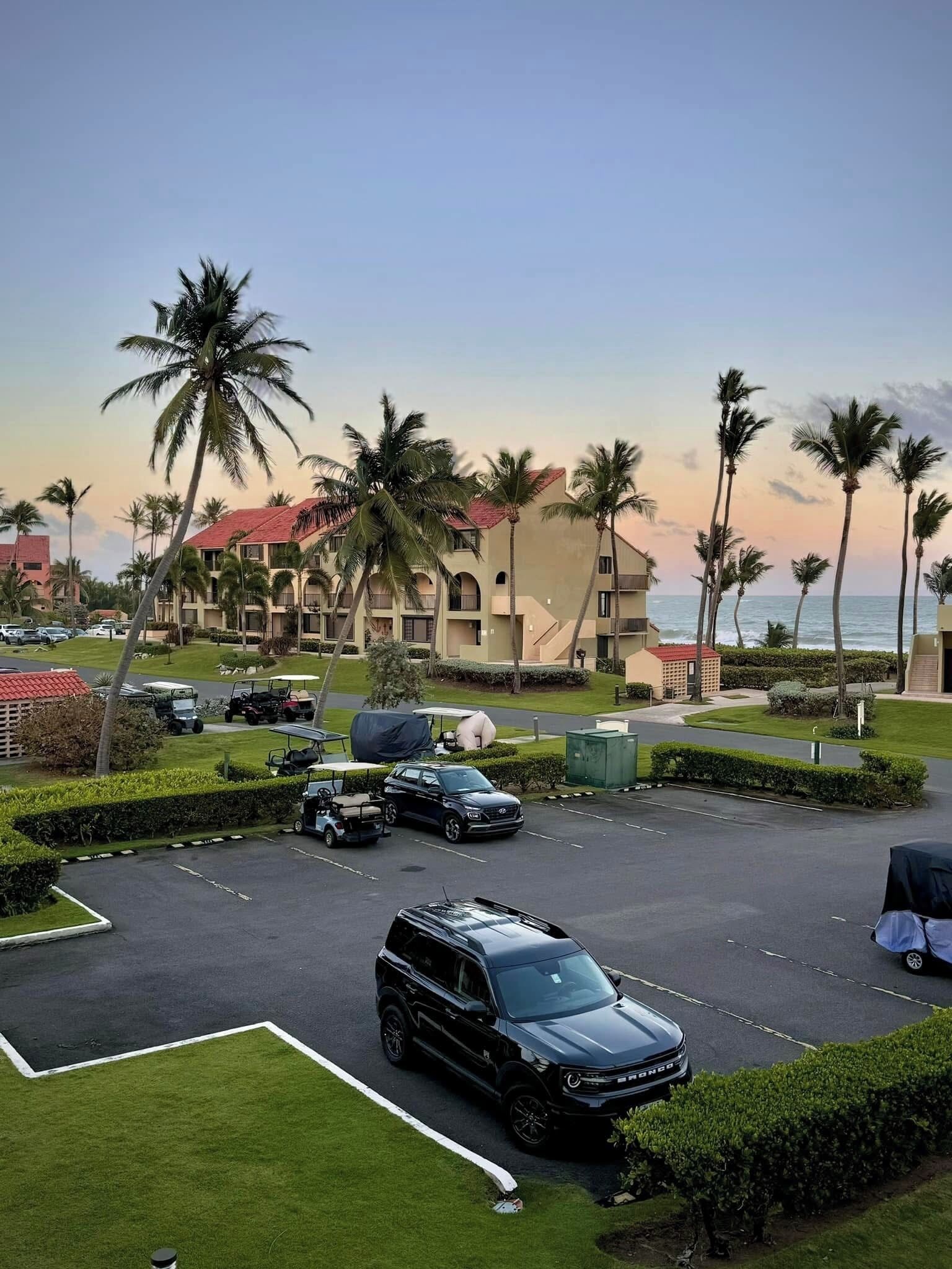 A parking lot with a black SUV, palm trees, and a building with a red roof in the background.