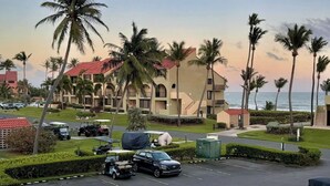 A parking lot with a black SUV, palm trees, and a building with a red roof in the background. - Carey Beach Villa at Palmas del Mar (Palmas del Mar)