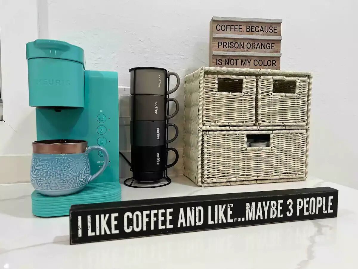A coffee maker, two coffee cups, a wicker basket, and a sign with a coffee-related message.