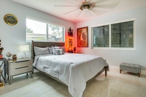 Une chambre à coucher comprenant un lit, des tables de chevet, une commode, un ventilateur de plafond, une fenêtre avec des stores et une horloge murale.