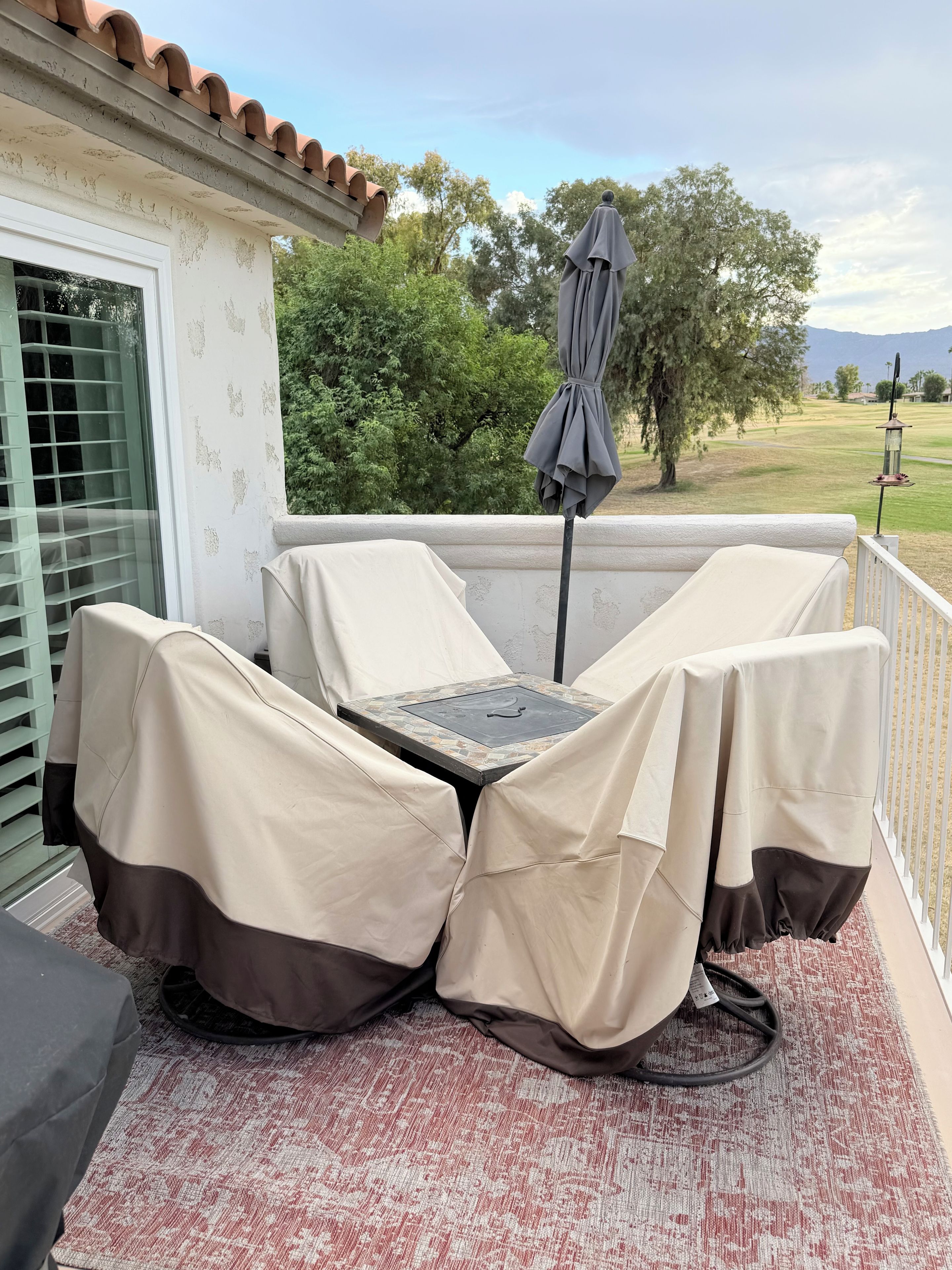 An outdoor patio with a covered seating area, a table with a metal top, and a view of a golf course and mountains in the background.