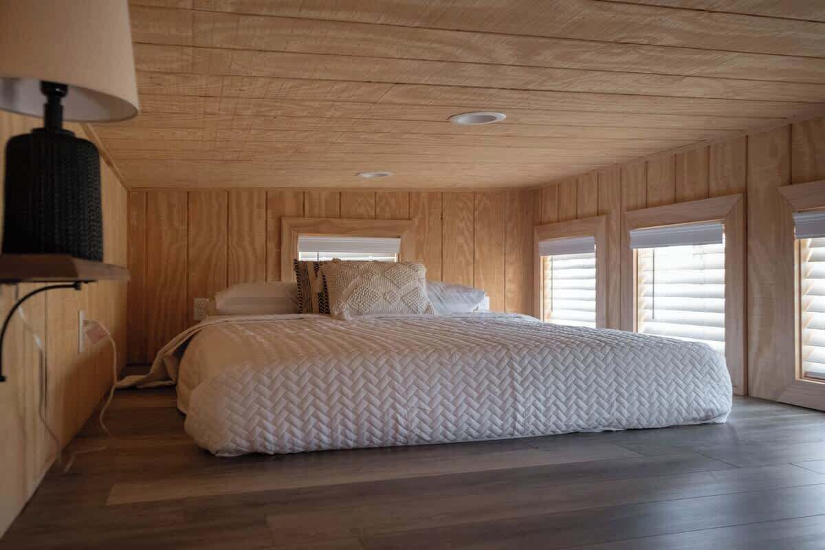 Une chambre à coucher avec un grand lit, des murs en bois, un plafond doté d’un éclairage encastré et une fenêtre avec des stores.