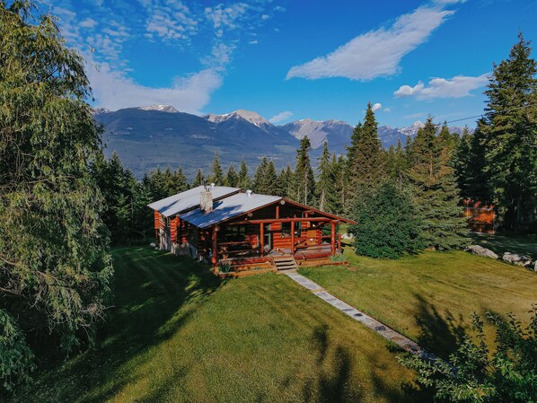 A log cabin with a stone chimney, surrounded by a well-maintained lawn and dense forest. - The Timberline Romance (Golden)