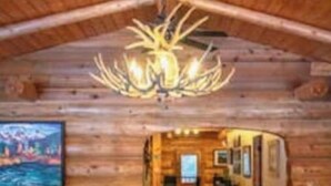 A log cabin interior with a wooden ceiling, a chandelier, a sofa, and a painting on the wall.