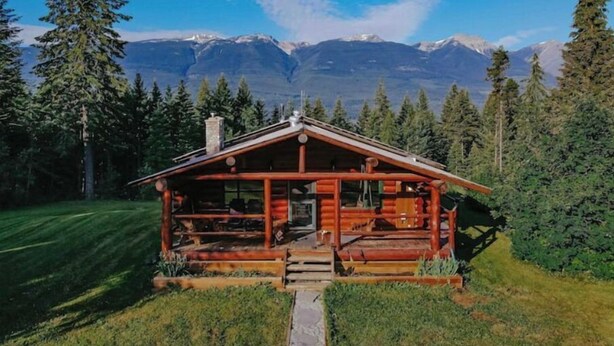 A log cabin with a porch, surrounded by a forest and mountains in the background.