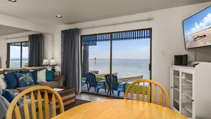 A room with a wooden table and chairs, a sofa with cushions, a TV mounted on the wall, and a view of the ocean through large sliding doors. - Las Brisas on the Bay Unit 119 (South Padre Island)
