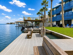 A wooden dock extending into a body of water with a pool area, palm trees, and a building with blue balconies.