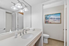 A bathroom with a white sink, a large mirror, and a toilet. There is a painting of Waikiki Beach on the wall. - 2nd Floor Condo Bayside, Windsurf, Fish Docks (South Padre Island)