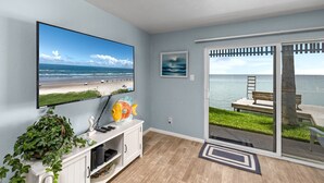 A room with a large TV displaying a beach scene, a small fish sculpture, a plant, and a door leading to an outdoor area with a bench and a view of the sea. - Las Brisas on the Bay Unit 116 (South Padre Island)