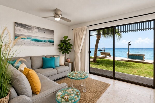 A living room with a sofa, a coffee table, a painting, and a view of the beach through large glass doors.