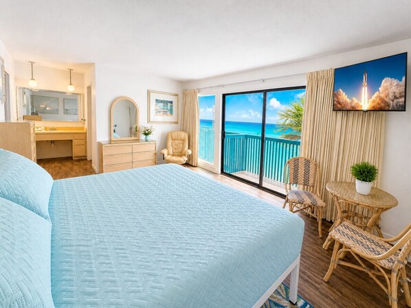 A hotel room with a large bed, a desk, a chair, a small table with chairs, a TV, and a balcony with a view of the ocean.