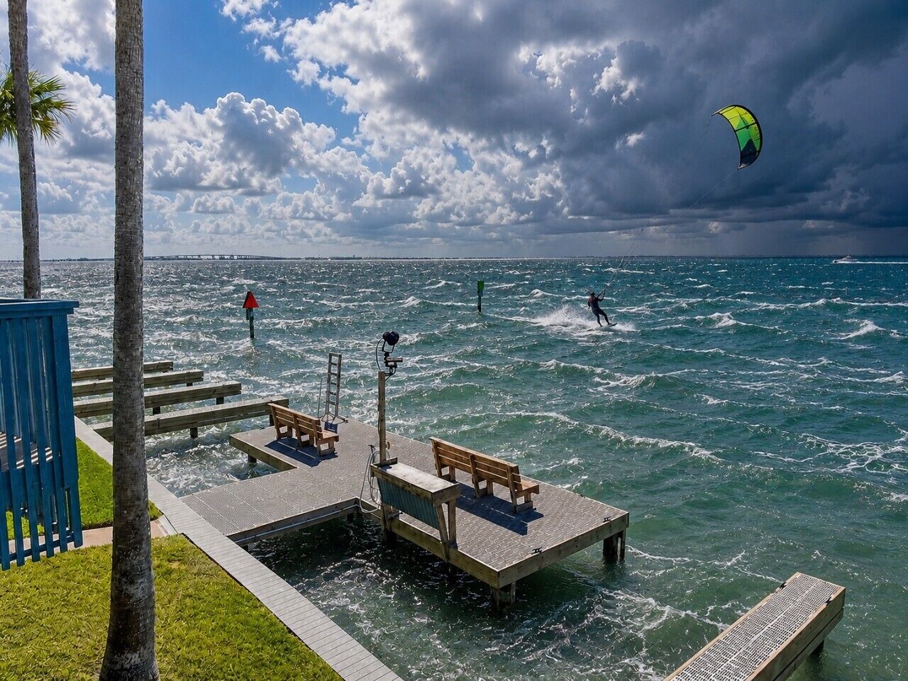 Een steiger met bankjes en een reddingsboei, uitkijkend over een wateroppervlak waar in de verte iemand aan het kitesurfen is.