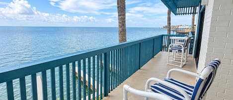 A balcony with two striped lounge chairs, a palm tree, and a view of the sea.