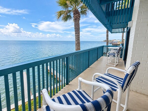 A balcony with two striped lounge chairs, a palm tree, and a view of the sea.