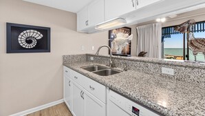 Una cocina con encimeras de granito, gabinetes blancos, un lavamanos de acero inoxidable y vista al océano a través de una ventana.