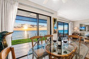 Une pièce avec une table en verre, des chaises, un canapé, un téléviseur et une vue sur l’océan au coucher du soleil.