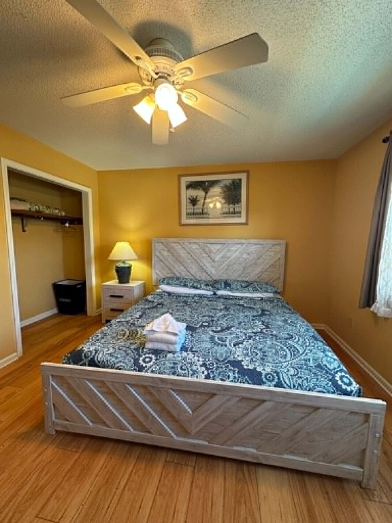 A bedroom with a bed, a ceiling fan, a nightstand, a lamp, a framed picture on the wall, and a closet.