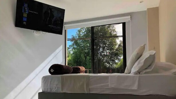 Una camera da letto con un letto, cuscini, una TV a parete e una finestra vista alberi.