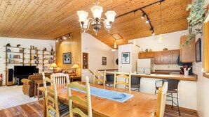 Amplia sala de estar con una mesa de comedor de madera, sillas, un sofá, un televisor, estanterías y una zona de cocina.