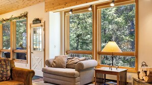 Un salón acogedor con un sofá, una mesa con lámpara y amplias ventanas con vistas a árboles.
