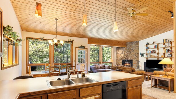 Una cocina amplia con armarios de madera, lavavajillas y una isla grande. En la sala de estar hay una chimenea de piedra, un televisor y una estantería.