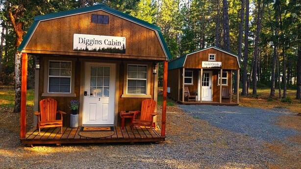 Zwei Holzhütten, genannt 'Diggins Cabin' und 'Gold Creek', mit einem Kiesweg dazwischen.